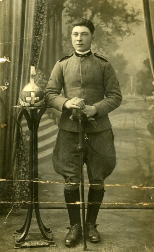 Militare in divisa in uno studio fotografico con sfrondo dipinto. Elmo poggiato su un portacappelli, guanti e una spada tra le mani, con la punta che poggia a terra